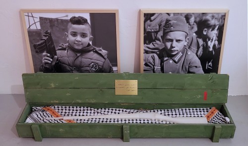 Krieg ist Frieden. Freiheit ist Sklaverei. Unwissenheit ist Stärke, Kastenobjekt mit Wachsgewehr und Kufiya, 2025